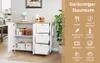 A white kitchen cart with a wooden countertop is shown in a kitchen setting. The cart has open shelves and drawers, and is filled with various kitchen items. A plant is visible to the right, and a wall with a framed picture is in the background. A section  Podgląd zdjęcia produktu