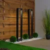 Two modern black bollard lights stand in a garden setting with a wooden fence backdrop, white gravel, and green spherical bushes. Produktbild-Vorschau