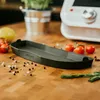 A dark gray kitchen accessory is placed on a wooden table with scattered peppercorns and salt, along with some tomatoes and a lemon in the background. A kitchen appliance is also visible. Náhľad obrázku produktu