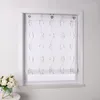 A white roller blind with a brown embroidered pattern is shown hanging in a window frame. A small bouquet of white flowers in a teal pot sits on a small table to the left of the window.