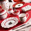 A collection of Christmas-themed tableware, including a vase, plates, sugar bowls, and a mug, arranged on a red tablecloth. Produktbild-Vorschau