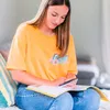 A young woman is sitting on a couch, looking down at a book and holding a device. Náhľad obrázku produktu