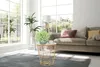 A living room scene with a sofa, large windows, and a coffee table with a plant and a cup of coffee. Produktbild-Vorschau