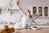 A woman is stretching in a kitchen with a kettle, mug, and pastries on the counter. Produktbild-Vorschau