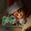 A child wearing a Santa hat sleeps peacefully next to a 3D illusion lamp shaped like a game controller. Produktbild-Vorschau