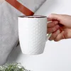 A hand holds a white ceramic mug with a honeycomb pattern, next to a grey fabric item with a brown leather strap. Podgląd zdjęcia produktu