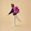 A young girl wearing a backpack walks against a plain background. Produktbild-Vorschau