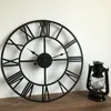 A large black metal wall clock with Roman numerals is displayed on a wooden surface against a white wall. A small black lantern is placed next to the clock. Produktbild-Vorschau