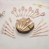 A collection of rose gold cutlery arranged on a light-colored surface, with a wooden block in the center holding some of the items. Podgląd zdjęcia produktu