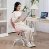 A woman is sitting on a pink office chair and reading a magazine in a well-lit room with a desk and a laptop. Produktbild-Vorschau