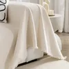 A cream-colored throw blanket draped over a sofa, with a decorative pillow visible on the left. A small side table with a candle is in the background. Produktbild-Vorschau