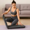 A woman in athletic wear is kneeling on a yoga mat, interacting with a piece of fitness equipment in a living room setting. Produktbild-Vorschau