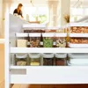 A woman is in the background of a kitchen, and in the foreground are several clear storage containers filled with various dry goods. Náhľad obrázku produktu