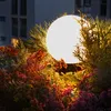 A close-up of a glowing spherical garden light surrounded by plants and flowers at dusk. Produktbild-Vorschau
