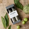 A gift box of salt and pepper spices is arranged on a wooden surface with fresh greens, an artichoke, and small green fruits. Produktbild-Vorschau