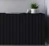A close-up view of a black cabinet with vertical slats, topped with a book, a small white bird figurine, and a potted plant. Produktbild-Vorschau