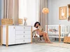 A woman reads a book in a nursery next to a crib and a dresser. Produktbild-Vorschau