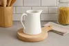 A white ceramic pitcher sits on a wooden cutting board on a kitchen counter, with a wooden utensil holder and a jar of pasta in the background. Produktbild-Vorschau