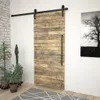 A wooden sliding barn door with a black handle and track, set against a white wall with a black and white tree artwork on the right and plants on the left. Produktbild-Vorschau