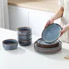 A person is holding a plate from a dinnerware set, with other plates and bowls stacked on a white countertop. Náhled obrázku produktu