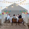 People dining under a tent on a beach at sunset, with string lights overhead. Voir les images du produit