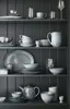 A collection of grey ceramic tableware displayed on shelves in a dark grey hutch. Produktbild-Vorschau