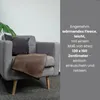 A brown fleece blanket draped over a gray armchair, with a green plant in the background and text overlay on the right. Produktbild-Vorschau