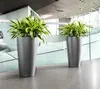 Two tall, gray planters with lush green and yellow plants are placed in a modern, well-lit interior space with glass walls and a concrete floor. Produktbild-Vorschau