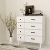 A white dresser with eight drawers, decorated with a vase of flowers, a framed picture, and a record player on top, placed in a room with a patterned rug on the floor. Produktbild-Vorschau