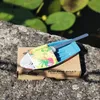 A small toy boat with a colorful sail is placed on a cardboard box, resting on a rock near a pond with lily pads. Produktbild-Vorschau