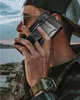 A person wearing a cap and glasses is holding a phone to their ear, with a rugged phone case on it. The background shows a body of water and rocks. Náhled obrázku produktu