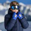 A young boy wearing ski goggles and a navy blue ski jacket, holding his gloved hands up to his face. The background is a blurred image of snowy mountains. Produktbild-Vorschau
