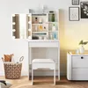 A white vanity table with a mirror and stool, along with a woven basket filled with pillows, is set against a white wall. Voir les images du produit