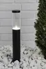 A black and clear bollard light is placed on white gravel in front of a white brick wall and a green bush. Produktbild-Vorschau