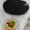 A black waffle maker is shown from a top-down perspective, with a finished waffle topped with salmon, greens, and roe on a plate in the foreground. Podgląd zdjęcia produktu