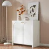 A white cabinet with arched doors is shown in a room with a floor lamp and artwork. Produktbild-Vorschau