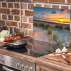 A kitchen scene with a stovetop, cooking utensils, and vegetables, featuring a glass splashback with a beach sunset image. Náhľad obrázku produktu