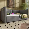 A modern gray upholstered daybed with integrated storage and charging ports, set in a well-lit room with a large window and artwork on the wall. Voir les images du produit