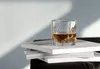 A glass of whiskey sits on a stack of books, with sunlight casting shadows on the wall. Produktbild-Vorschau