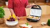 A person is using a sandwich maker in a kitchen setting, with fruit and other food items visible. Podgląd zdjęcia produktu