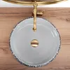 A top-down view of a round ceramic sink with a decorative blue and white rim, a gold faucet, and a gold drain cover, placed on a wooden surface. Produktbild-Vorschau
