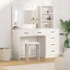 A white vanity table with a mirror and shelves, along with a stool, is set up in a room with a plant and basket. Voir les images du produit