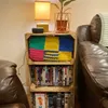 A wooden crate shelf filled with DVDs and a colorful knitted blanket, next to a leather couch and a lamp. Produktbild-Vorschau