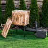 A wooden smoker oven with a detached side drawer, set up on a green lawn in front of a wooden fence and evergreen trees. Náhled obrázku produktu