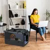 A woman is working on her laptop while sitting in a chair next to a dog carrier with a dog inside. Anteprima dell’immagine prodotto