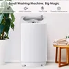 A small white washing machine is shown in a brightly lit room with a plant and a stool with towels. Produktbild-Vorschau