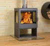 A modern wood-burning stove with a glass door and a visible fire is placed on a glass hearth in front of a stone-tiled wall. Produktbild-Vorschau