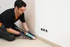 A man is kneeling and applying sealant to the base of a wall with a caulking gun. Produktbild-Vorschau