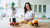 A woman is preparing fruit in a kitchen with a blender and a bowl of fruit on the counter. Podgląd zdjęcia produktu