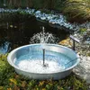 A solar-powered fountain kit is operating in a galvanized metal basin filled with water, placed outdoors among plants and rocks near a pond. Produktbild-Vorschau